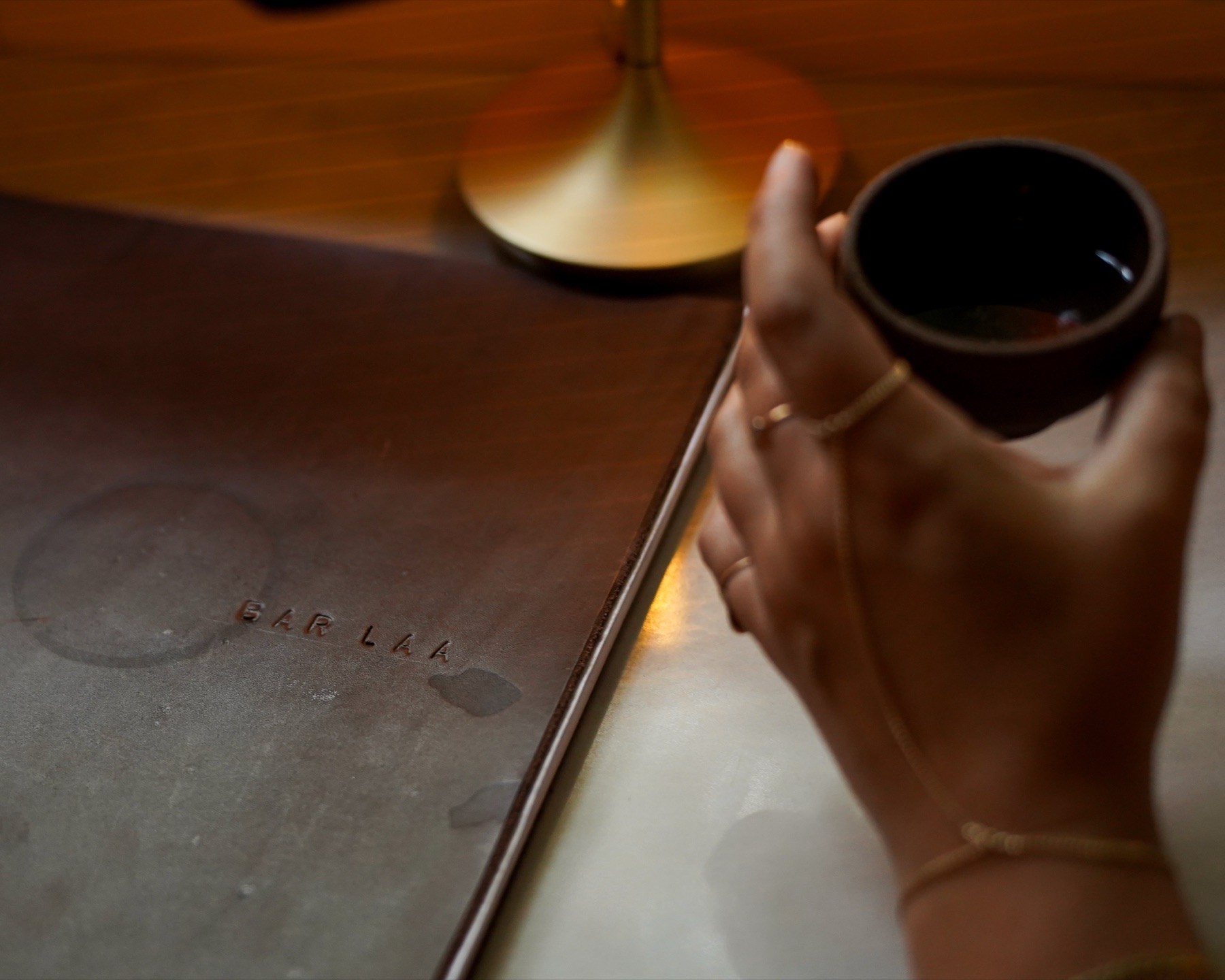 A hand holds a small clay copita beside a leather Bar Laa menu, candlelit